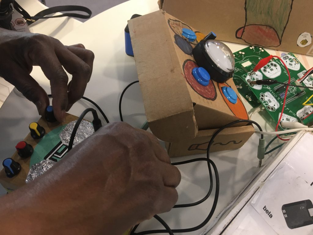 The hands of a visitor to the exhibit turn dials on a cardboard box with an embedded set of dials, cables and arcade buttons.